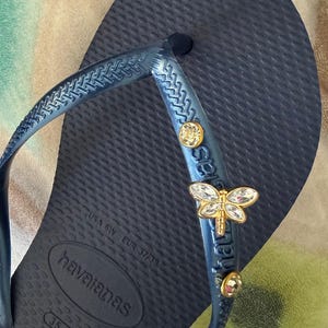 May include: Navy blue Havaianas flip-flop with a textured footbed. The strap is adorned with a gold-colored dragonfly charm and two round, clear crystal accents. The flip-flop is a size USA 6W, EUR 37/38.