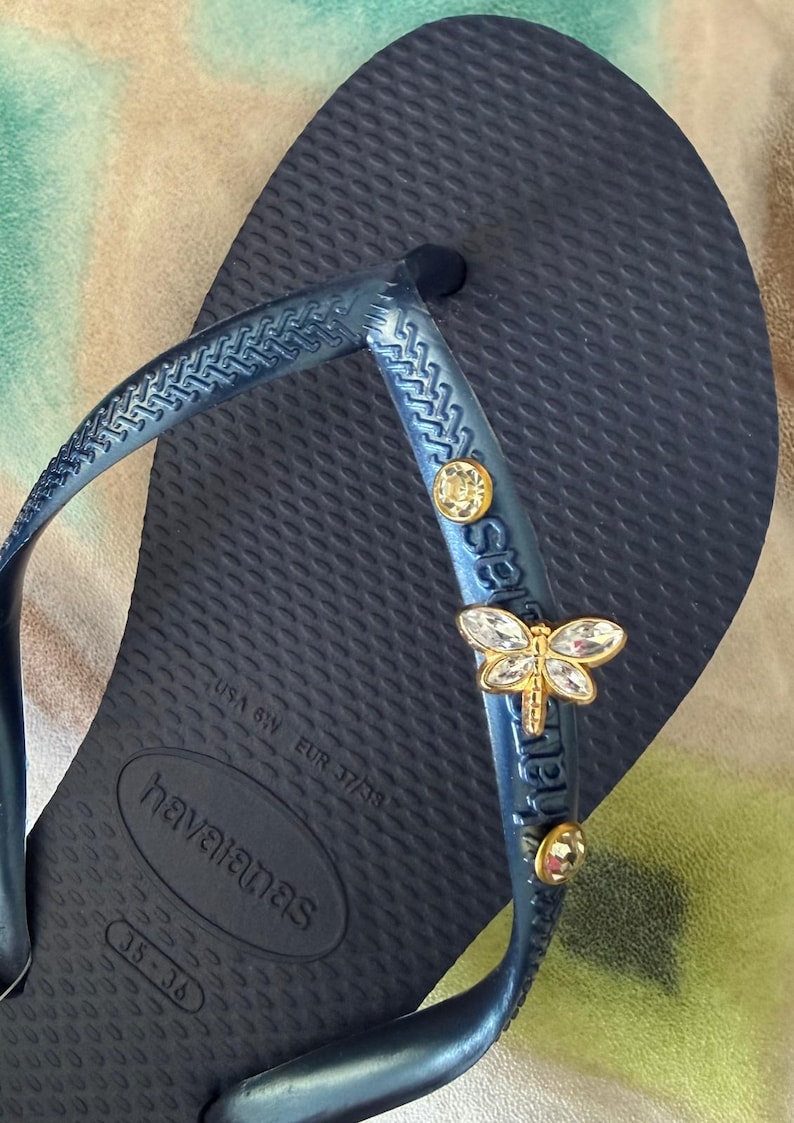 May include: Navy blue Havaianas flip-flop with a textured footbed. The strap is adorned with a gold-colored dragonfly charm and two round, clear crystal accents. The flip-flop is a size USA 6W, EUR 37/38.