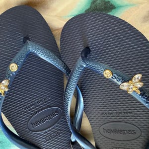 May include: Navy blue Havaianas flip-flops with decorative gold and crystal bee and gem accents. The flip-flops have a textured footbed and a blue strap. The brand name "Havaianas" is visible on the footbed and strap.