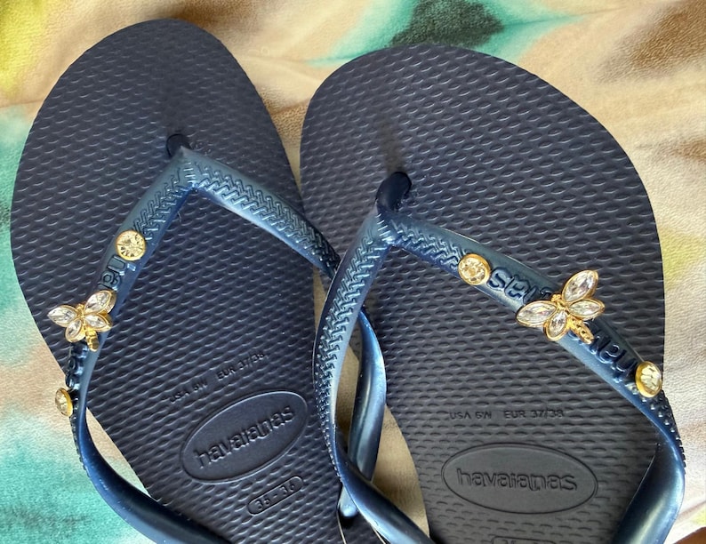 May include: Navy blue Havaianas flip-flops with decorative gold and crystal bee and gem accents. The flip-flops have a textured footbed and a blue strap. The brand name "Havaianas" is visible on the footbed and strap.