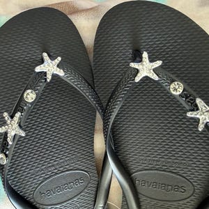 K&ouml;nnte beinhalten: Schwarze Havaianas-Flip-Flops mit Strass-Seesternen und Edelsteinverzierungen. Die Sandalen haben ein strukturiertes Fu&szlig;bett und schwarze Riemen. Das Havaianas-Logo ist auf der Sohle sichtbar. Eine stilvolle und strandtaugliche Schuhoption.