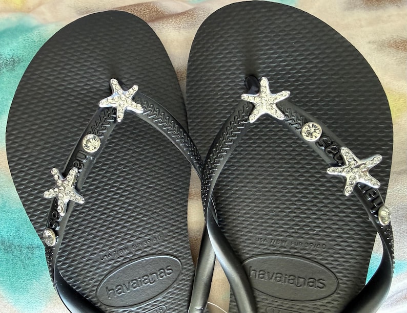 K&ouml;nnte beinhalten: Schwarze Havaianas-Flip-Flops mit Strass-Seesternen und Edelsteinverzierungen. Die Sandalen haben ein strukturiertes Fu&szlig;bett und schwarze Riemen. Das Havaianas-Logo ist auf der Sohle sichtbar. Eine stilvolle und strandtaugliche Schuhoption.