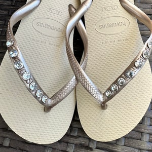 May include: A pair of beige flip-flops with gold straps and rhinestones. The straps are decorated with the word "Havaianas" and the size "US 7/38 EUR 38/39".