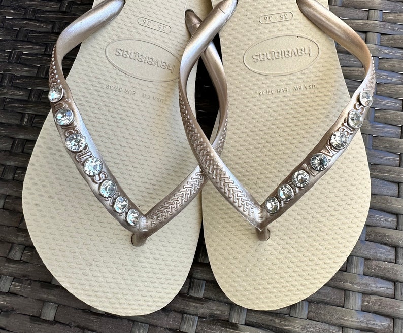 May include: A pair of beige flip-flops with gold straps and rhinestones. The straps are decorated with the word "Havaianas" and the size "US 7/38 EUR 38/39".
