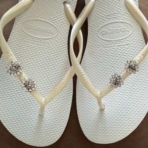 以下が含まれることがあります： 白い Havaianas ビーチサンダルのペア。ストラップには装飾が施されています。各ストラップには、透明なクリスタルアクセントが付いた、銀色の花型の装飾が2つ付いています。ビーチサンダルはテクスチャード加工されたフットベッドを備えています。