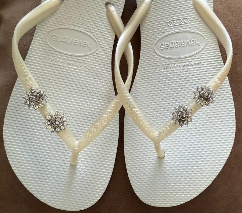 以下が含まれることがあります： 白い Havaianas ビーチサンダルのペア。ストラップには装飾が施されています。各ストラップには、透明なクリスタルアクセントが付いた、銀色の花型の装飾が2つ付いています。ビーチサンダルはテクスチャード加工されたフットベッドを備えています。