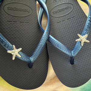 K&ouml;nnte beinhalten: Ein Paar marineblaue Havaianas-Flip-Flops mit blauen Riemen. Jeder Riemen ist mit einem silbernen Seestern verziert. Die Flip-Flops sind in der EU-Gr&ouml;&szlig;e 39/40 erh&auml;ltlich. Das Havaianas-Logo ist sichtbar.