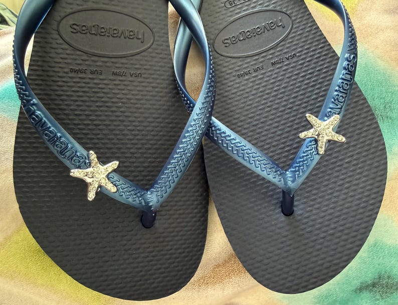 K&ouml;nnte beinhalten: Ein Paar marineblaue Havaianas-Flip-Flops mit blauen Riemen. Jeder Riemen ist mit einem silbernen Seestern verziert. Die Flip-Flops sind in der EU-Gr&ouml;&szlig;e 39/40 erh&auml;ltlich. Das Havaianas-Logo ist sichtbar.