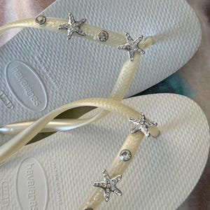 K&ouml;nnte beinhalten: Wei&szlig;e Havaianas-Flip-Flops mit strukturierter Sohle. Die Riemen sind mit silbernen Seesternen und klaren Strasssteinen verziert. Die Flip-Flops sind Gr&ouml;&szlig;e 37-38, was etwa 23 cm entspricht.