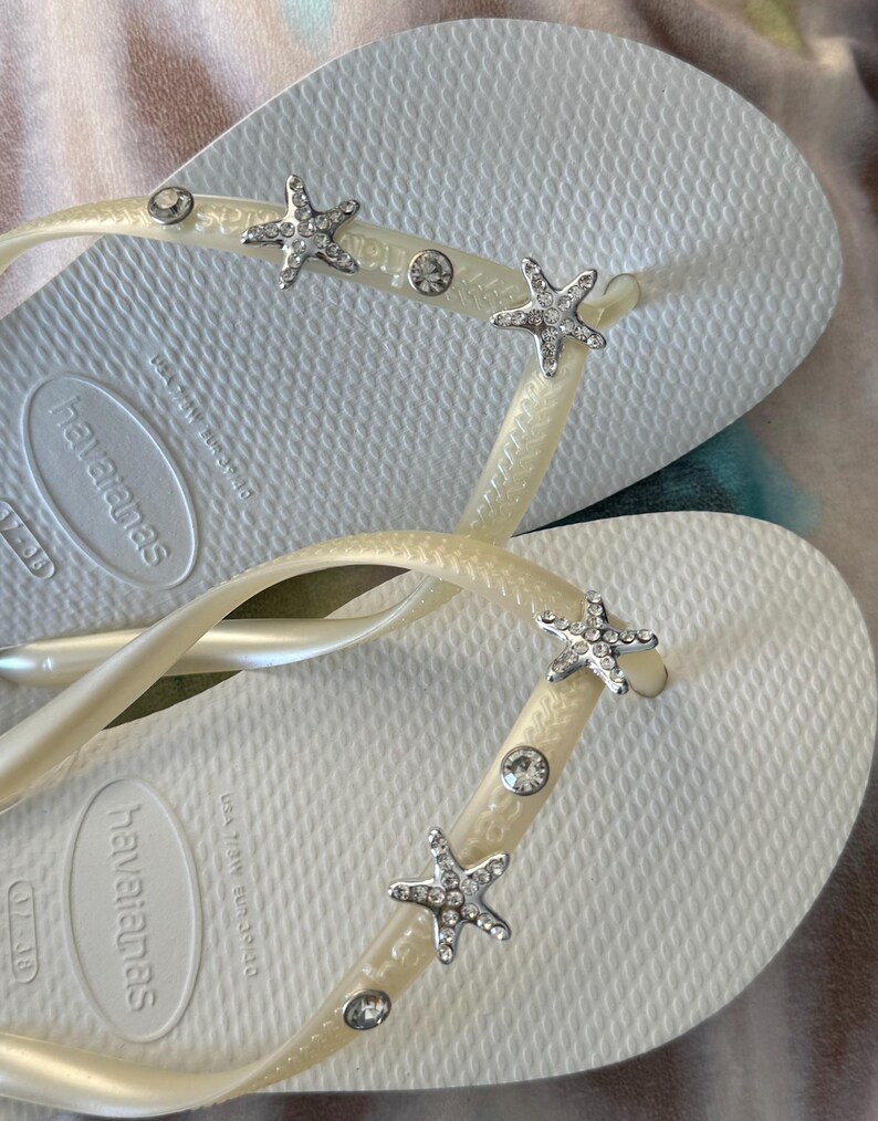 K&ouml;nnte beinhalten: Wei&szlig;e Havaianas-Flip-Flops mit strukturierter Sohle. Die Riemen sind mit silbernen Seesternen und klaren Strasssteinen verziert. Die Flip-Flops sind Gr&ouml;&szlig;e 37-38, was etwa 23 cm entspricht.