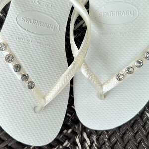 May include: White flip flops with a rhinestone embellishment on the strap. The flip flops have the brand name "Havaianas" printed on the sole.
