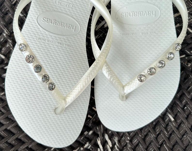 May include: White flip flops with a rhinestone embellishment on the strap. The flip flops have the brand name "Havaianas" printed on the sole.
