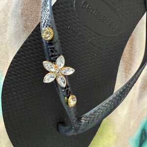 May include: Black Havaianas flip-flop with a decorative flower and gold-colored embellishments. The flower is made of clear crystals and gold-colored metal. The flip-flop has a textured footbed and straps.