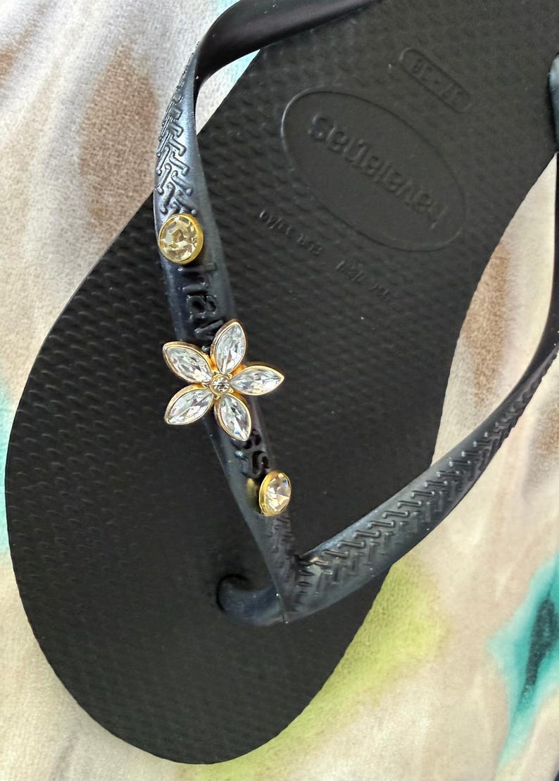 May include: Black Havaianas flip-flop with a decorative flower and gold-colored embellishments. The flower is made of clear crystals and gold-colored metal. The flip-flop has a textured footbed and straps.