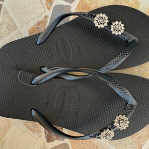 以下が含まれることがあります： 装飾的なシルバーの花のアクセントが付いた黒いビーチサンダル。サンダルは、テクスチャード加工されたフットベッドと黒いストラップを備えています。花のアクセントは、小さな輝く石で作られています。ビーチサンダルはカジュアルな履物です。