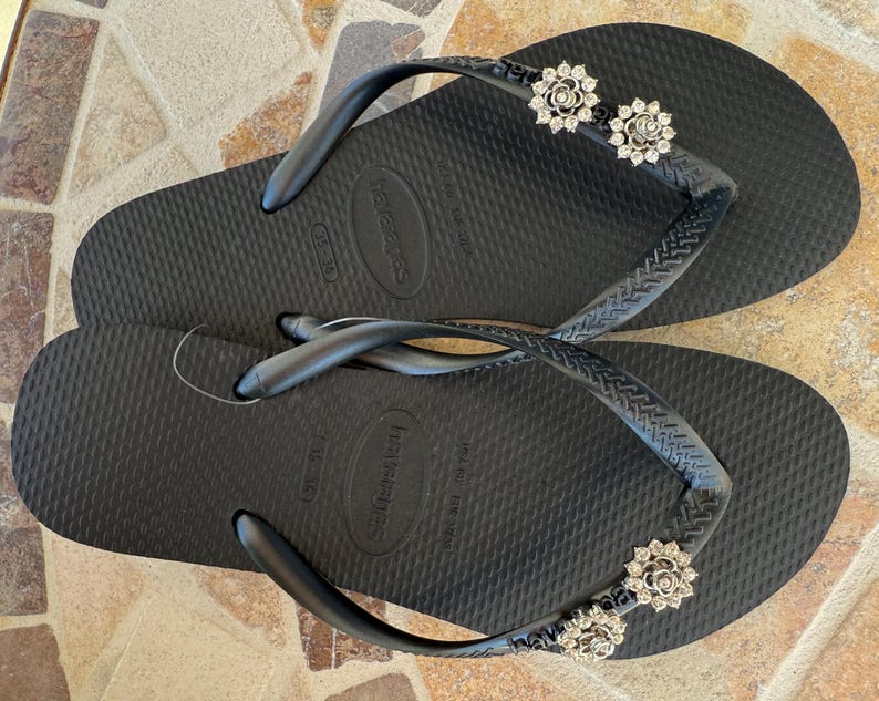 以下が含まれることがあります： 装飾的なシルバーの花のアクセントが付いた黒いビーチサンダル。サンダルは、テクスチャード加工されたフットベッドと黒いストラップを備えています。花のアクセントは、小さな輝く石で作られています。ビーチサンダルはカジュアルな履物です。