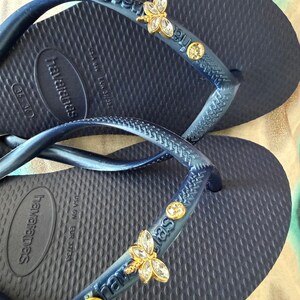 May include: Navy blue Havaianas flip-flops with embellished straps. The straps feature gold-toned butterfly charms and clear, round gemstones. The flip-flops have a textured footbed and the Havaianas logo.