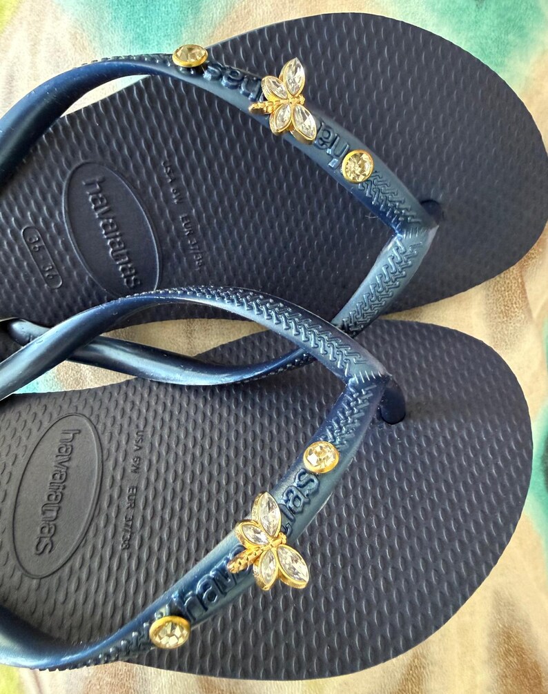 May include: Navy blue Havaianas flip-flops with embellished straps. The straps feature gold-toned butterfly charms and clear, round gemstones. The flip-flops have a textured footbed and the Havaianas logo.