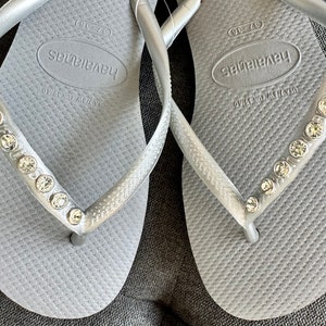 May include: A pair of silver flip-flops with rhinestones on the straps. The flip-flops have the brand name "Havaianas" printed on the footbed.
