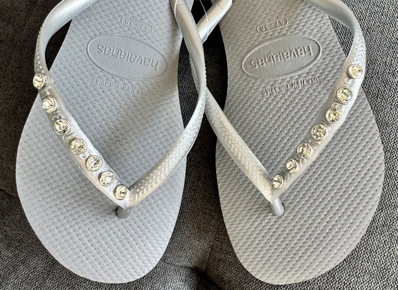 May include: A pair of silver flip-flops with rhinestones on the straps. The flip-flops have the brand name "Havaianas" printed on the footbed.
