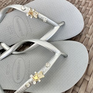 Puede incluir: Un par de chanclas plateadas con un adorno dorado y de cristal en cada tira. Las chanclas tienen el texto "havaianas" impreso en la plantilla. La talla es 37-38.