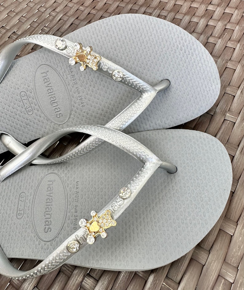 Puede incluir: Un par de chanclas plateadas con un adorno dorado y de cristal en cada tira. Las chanclas tienen el texto "havaianas" impreso en la plantilla. La talla es 37-38.