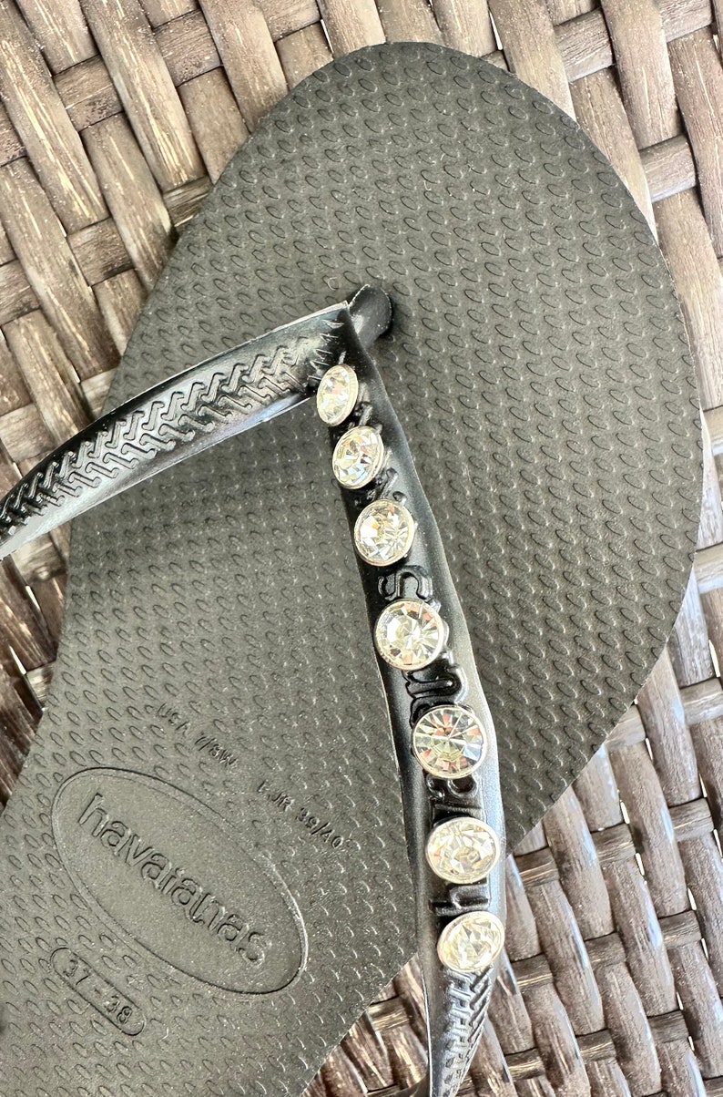 May include: Black flip-flops with a row of rhinestones on the strap. The flip-flops have the text "havaianas" and "37-38" printed on the sole.