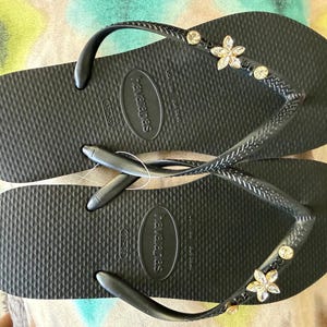 May include: Black flip-flops with a textured surface. The straps are embellished with small, clear gemstones and flower-shaped decorations. The brand name "havaianas" is visible on the sole.