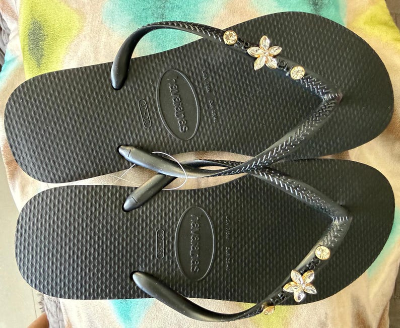May include: Black flip-flops with a textured surface. The straps are embellished with small, clear gemstones and flower-shaped decorations. The brand name "havaianas" is visible on the sole.