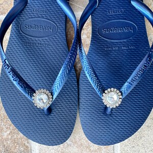 May include: Navy blue Havaianas flip-flops, size 37-38, with a clear rhinestone embellishment. The flip-flops feature the Havaianas logo on the strap and footbed. The size is equivalent to a UK size 4-5.