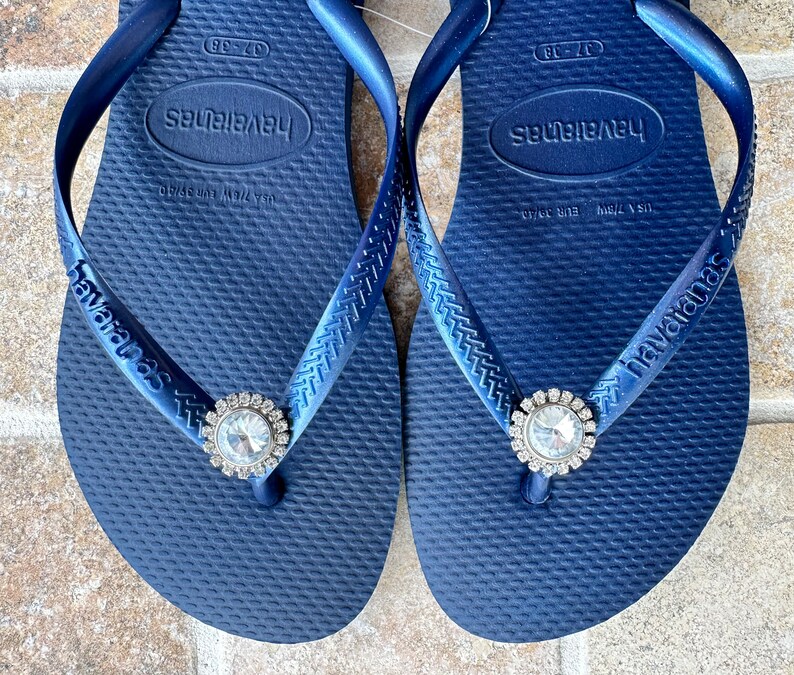 May include: Navy blue Havaianas flip-flops, size 37-38, with a clear rhinestone embellishment. The flip-flops feature the Havaianas logo on the strap and footbed. The size is equivalent to a UK size 4-5.