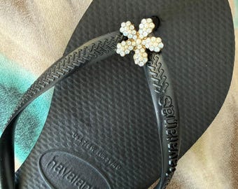 May include: A black Havaianas flip-flop featuring a textured footbed and a Y-shaped strap. The strap is adorned with a decorative, six-petaled flower charm encrusted with small, clear, sparkling rhinestones at the center junction.