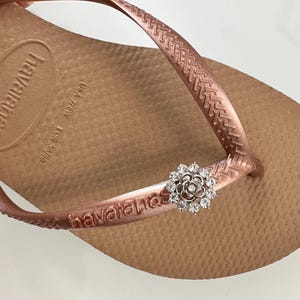 Pode incluir: Havaianas cor ouro rosa com um charme de flor brilhante. As Havaianas t&ecirc;m a palavra "havaianas" impressa na tira.
