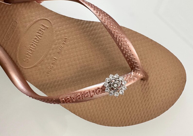 Pode incluir: Havaianas cor ouro rosa com um charme de flor brilhante. As Havaianas t&ecirc;m a palavra "havaianas" impressa na tira.