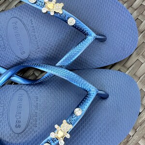 Puede incluir: Un par de chanclas azules con un dije de oso dorado y diamantes de imitaci&oacute;n en la correa. Las chanclas son de la marca Havaianas y tienen la informaci&oacute;n de talla "USA 7/8W EUR 39/40" impresa en la suela.