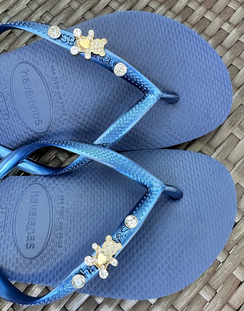 Puede incluir: Un par de chanclas azules con un dije de oso dorado y diamantes de imitaci&oacute;n en la correa. Las chanclas son de la marca Havaianas y tienen la informaci&oacute;n de talla "USA 7/8W EUR 39/40" impresa en la suela.