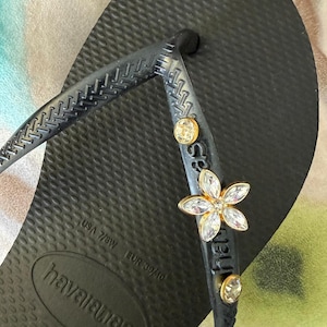 May include: Black Havaianas flip-flop with a textured sole. The strap is embellished with a gold-toned flower and two round, clear gemstones. The flip-flop is a USA size 7/8W.