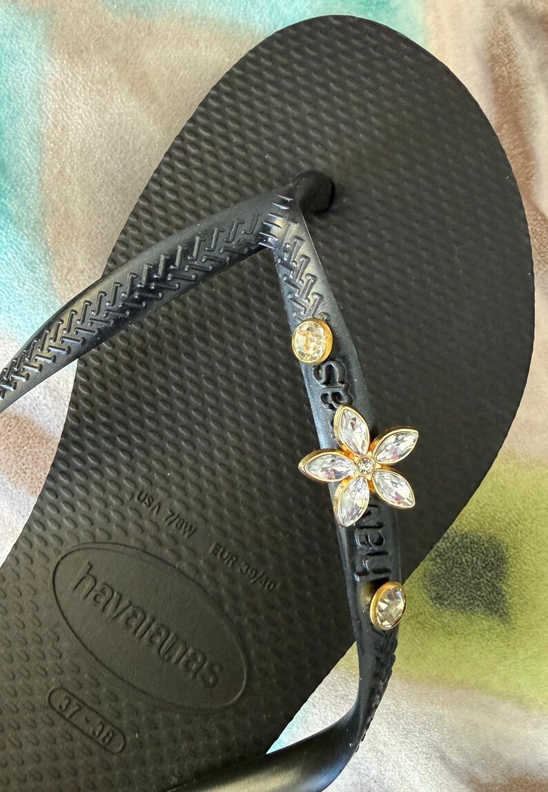 May include: Black Havaianas flip-flop with a textured sole. The strap is embellished with a gold-toned flower and two round, clear gemstones. The flip-flop is a USA size 7/8W.
