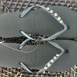 May include: A pair of black flip-flops with a silver strap and rhinestones. The flip-flops have the brand name "havaianas" printed on the sole.
