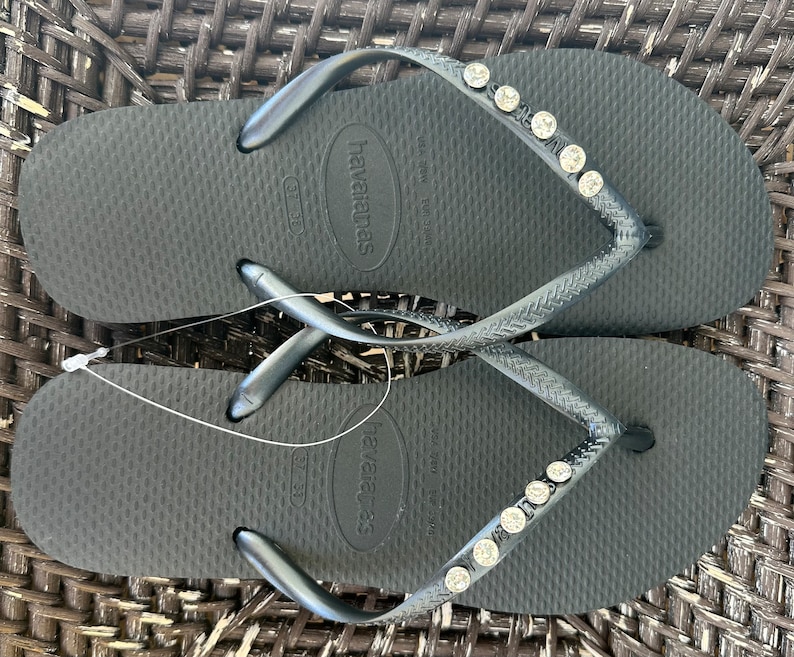 May include: A pair of black flip-flops with a silver strap and rhinestones. The flip-flops have the brand name "havaianas" printed on the sole.
