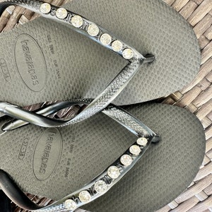 May include: Black flip-flops with a row of rhinestones on the strap. The flip-flops have the text "havaianas" and "USA 7/38 EUR 38/39" printed on the sole.