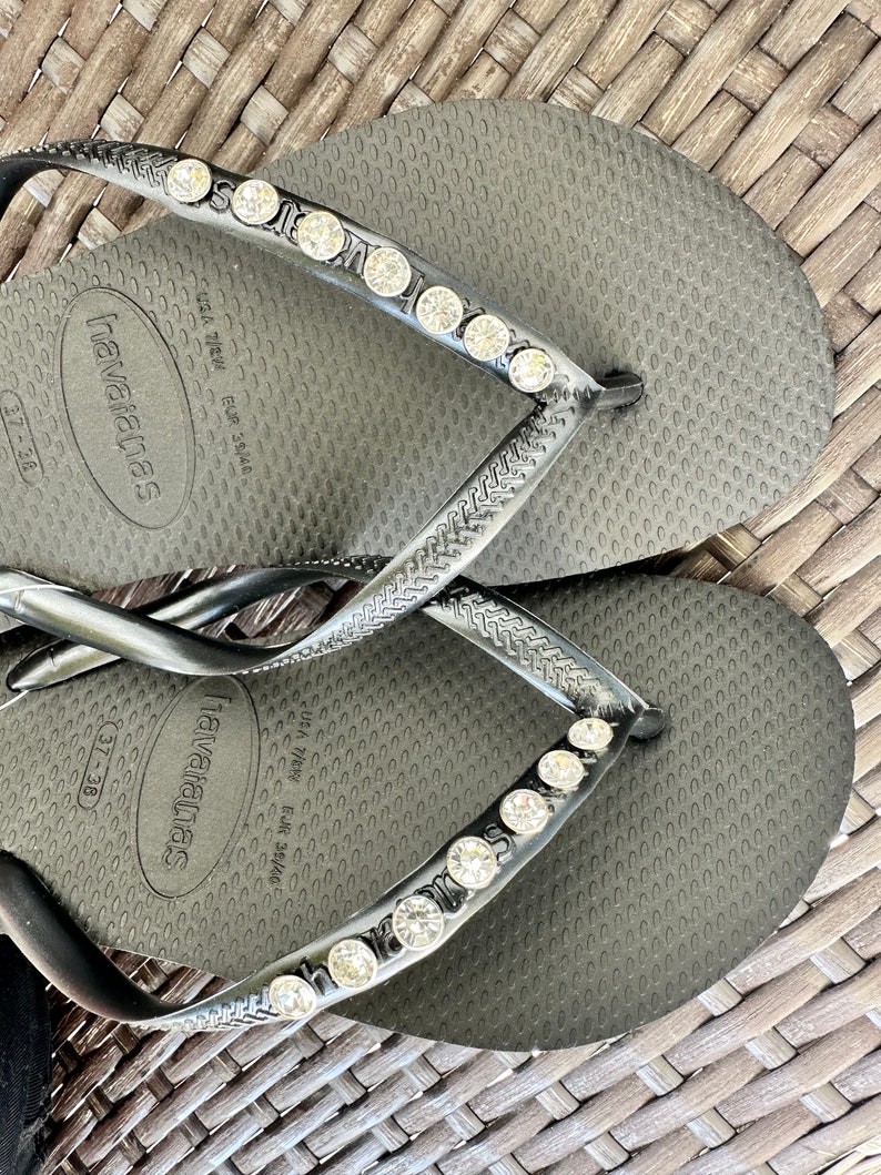 May include: Black flip-flops with a row of rhinestones on the strap. The flip-flops have the text "havaianas" and "USA 7/38 EUR 38/39" printed on the sole.