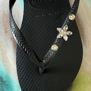May include: Black Havaianas flip-flop with a textured footbed and strap. The strap is embellished with a flower-shaped jewel and two round jewels. The flip-flop is a size 7/8 US, EUR 39/40.