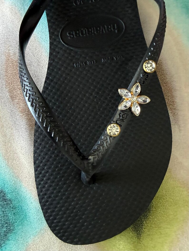 May include: Black Havaianas flip-flop with a textured footbed and strap. The strap is embellished with a flower-shaped jewel and two round jewels. The flip-flop is a size 7/8 US, EUR 39/40.
