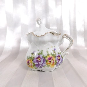 Pode incluir: Uma açucareiro de porcelana branca com tampa e alça. A tigela é decorada com amores-perfeitos roxos, amarelos e brancos. A borda e a alça têm acabamento dourado. A tigela está em um fundo branco listrado.