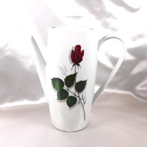 Puede incluir: Cafetera de cerámica blanca con un pico largo y asa. Una rosa roja con hojas verdes está pintada en la parte delantera. La cafetera está sobre un fondo blanco.