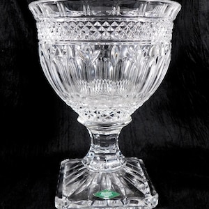 May include: A clear, cut-glass bowl with a wide, flared rim and a pedestal base. The bowl features intricate diamond and leaf patterns, and sits on a square base. The overall design is elegant and decorative.