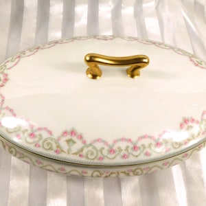 Puede incluir: Fuente ovalada blanca con tapa, con asas doradas y un borde floral decorativo rosa y verde. El plato está diseñado para servir alimentos y tiene un asa dorada en la tapa.