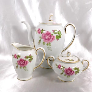Pode incluir: Um conjunto de chá de porcelana branca com bule, cremeira e açucareiro. Cada peça é adornada com desenhos florais de rosas cor-de-rosa e detalhes dourados. O bule tem um bico e uma alça curvos, e o açucareiro tem uma tampa.