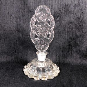 May include: A clear glass perfume bottle featuring a wide, circular base with vertical fluting and a decorative, tall stopper. The stopper is composed of three stacked, circular medallions, each intricately embossed with a starburst pattern.